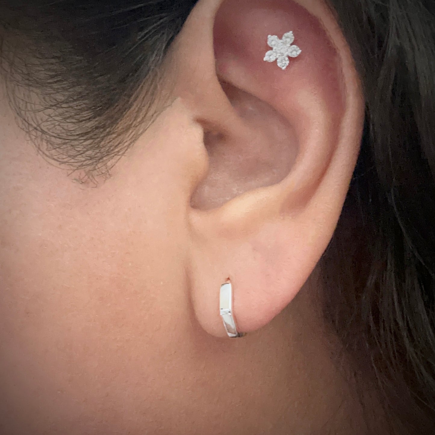 Model wearing a sterling silver hexagonal-shaped huggie hoop earring. The earring is sitting snug against her lobe. The model is also wearing a flower-shaped white cubic zirconia stud in the flat part of her ear just under the upper helix.