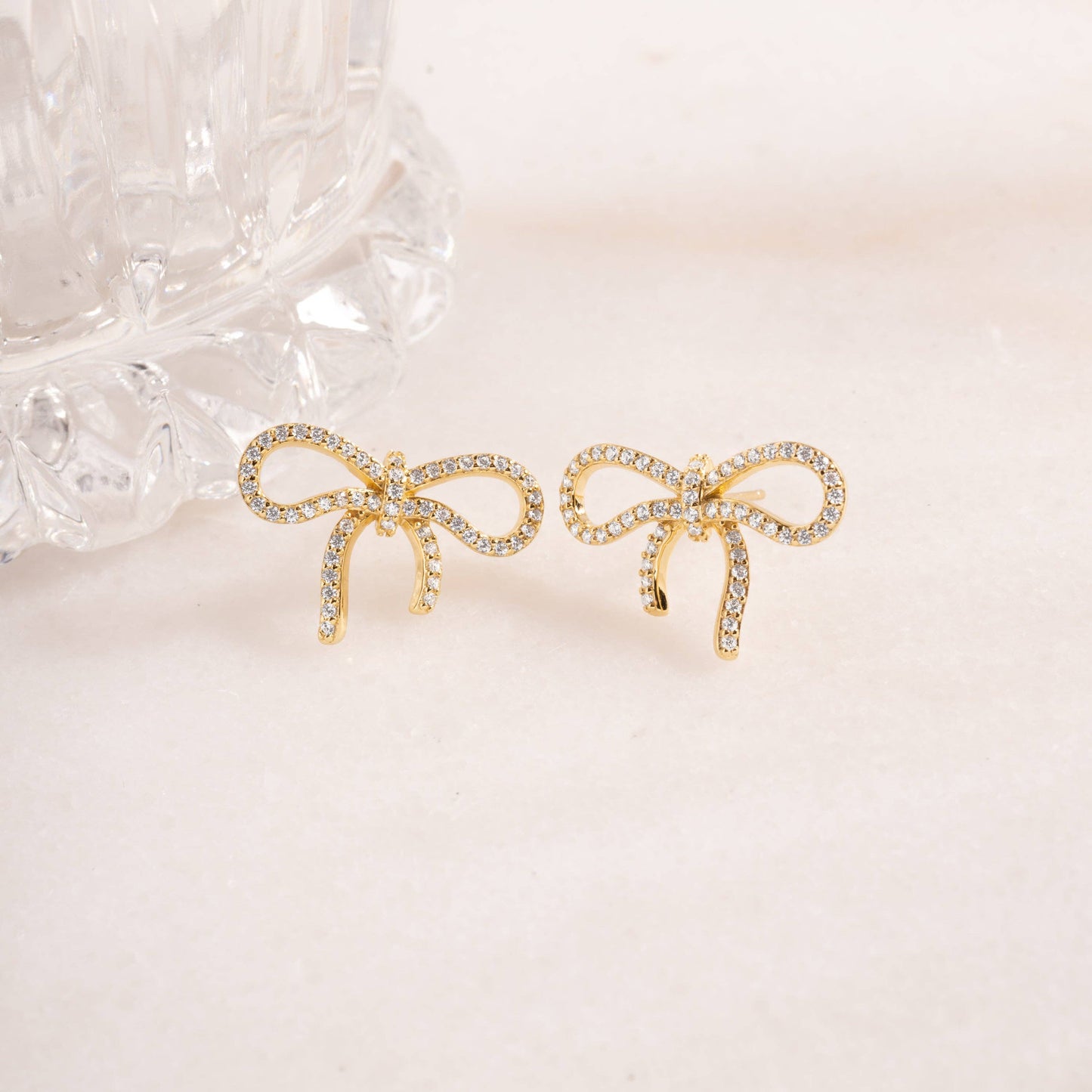 Gold-plated sterling silver stud earrings in the shape of a bow against a light pink background. Each bow is lined with pave-set white cubic zirconia.