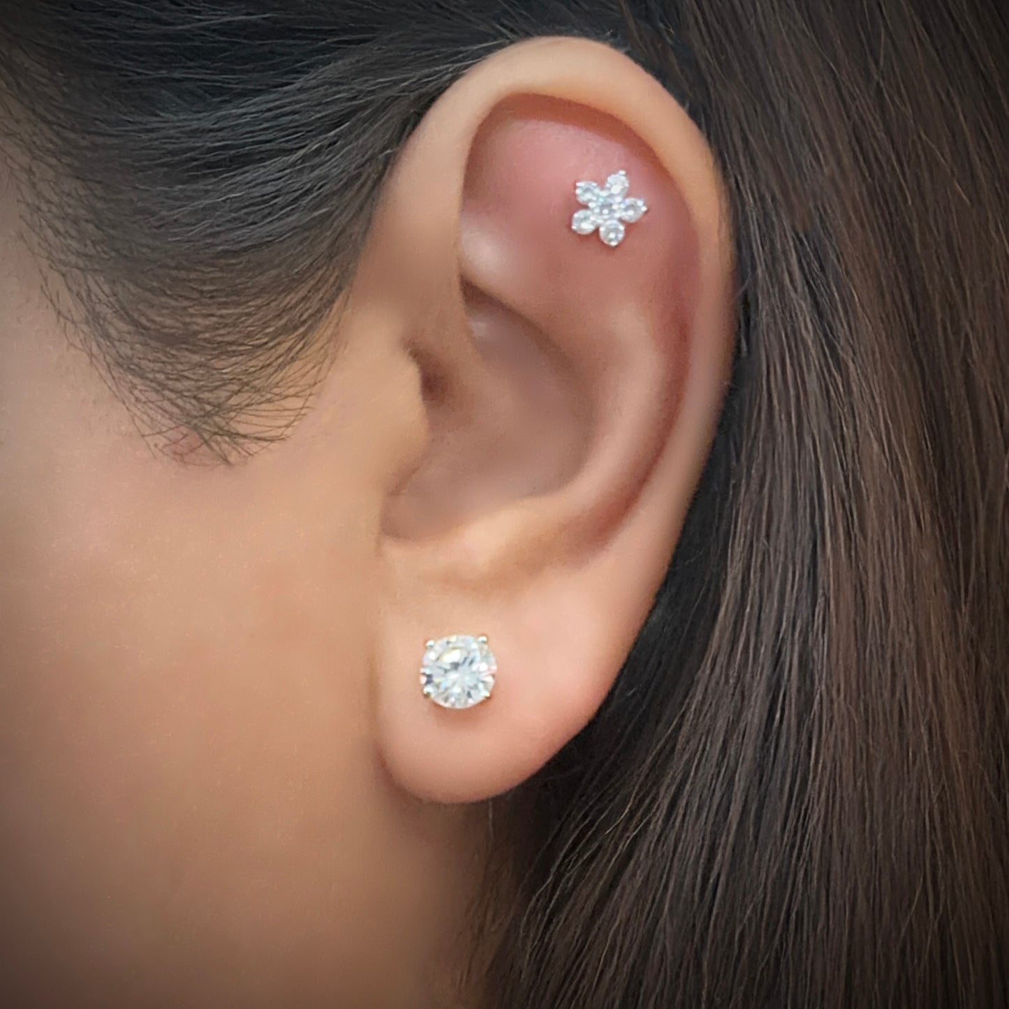 Model wearing a single solitaire white cubic zirconia stud earring in sterling silver. The model also has a flower-shaped white cubic stud earring on the flat portion of her ear just below the upper helix.
