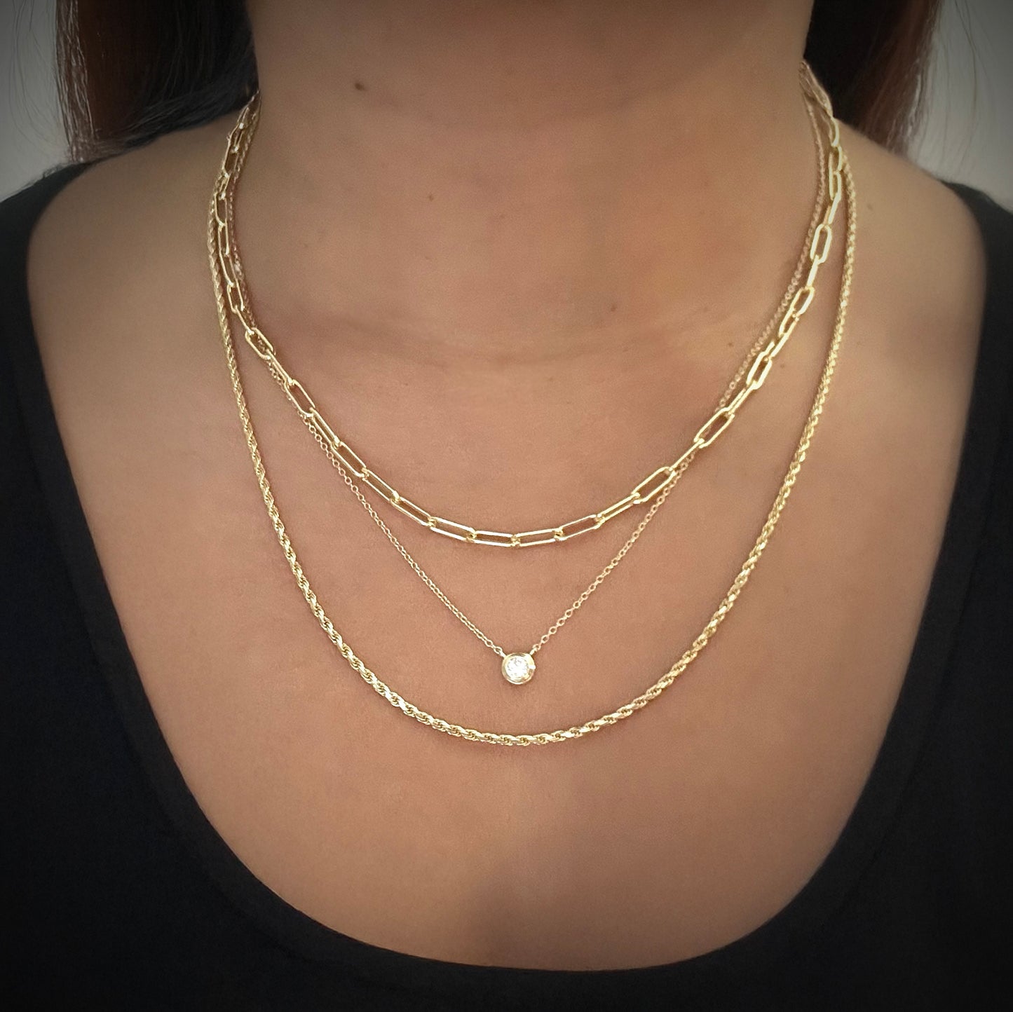 Model wearing a gold-plated sterling silver delicate chain necklace with bezel set white cubic zirconia pendant connected to the chain. The model has layered this necklace with 2 other gold-plated silver necklaces; one is a paperclip style necklace and the other a rope chain necklace.