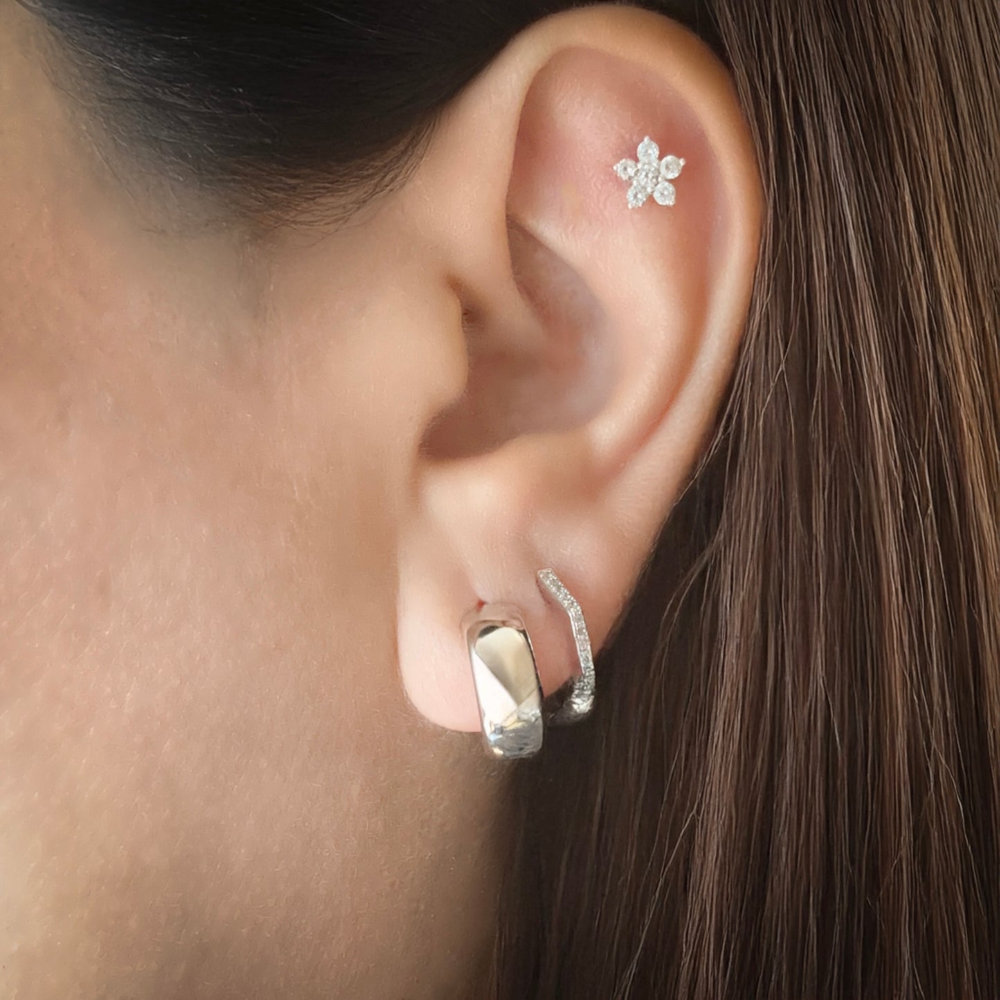 A model wearing a sterling silver chunky huggie hoop earring in her first lobe piercing. She has a hexagonal-shaped huggie lined with white cubic zirconia in her second lobe piercing and a flower-shaped white cubic zirconia stud in the flat part of her ear just below the upper helix.