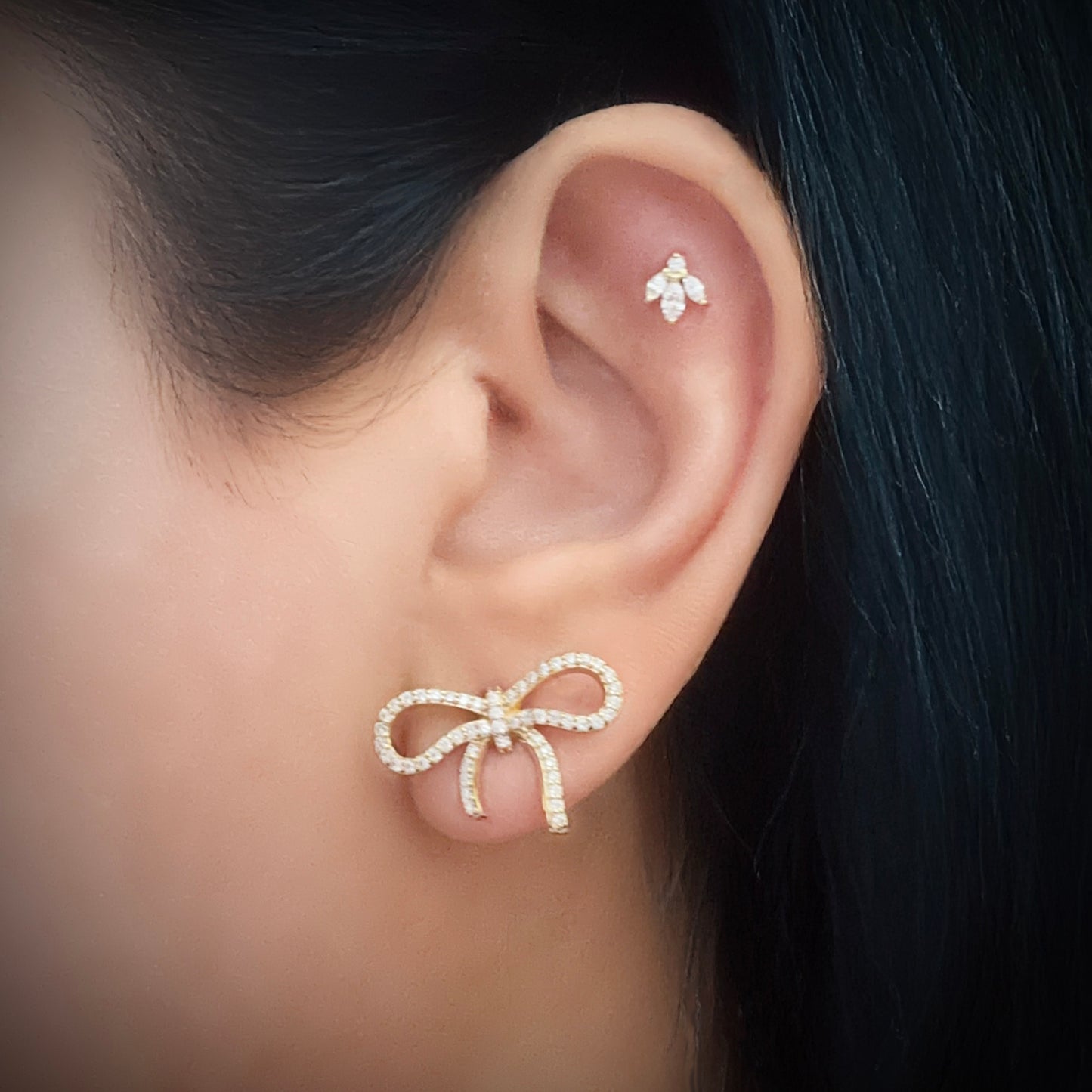 Model wearing a gold-plated sterling silver stud earring in the shape of a bow. The bow is lined with pave-set white cubic zirconia. The model is also wearing a flower-shaped white cubic zirconia stud with three stone petals sitting below a center stone in the flat part of her ear just under the upper helix.