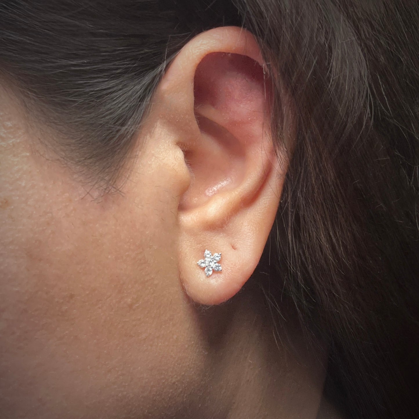 Model wearing a sterling silver flower-shaped stud earring. The earring has 5 petals made of white cubic zirconia surrounding a center stone also made of white cubic zirconia.