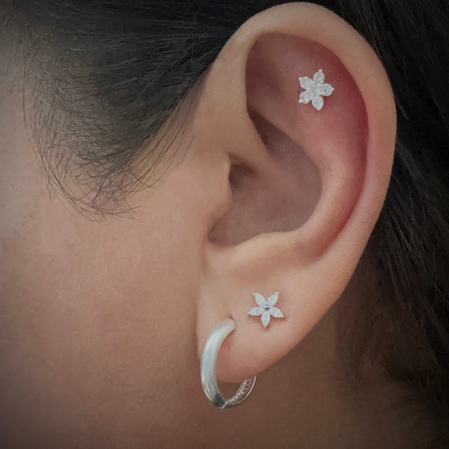 Model wearing a slightly chunky sterling silver hoop in her first lobe piercing. In her second piercing, she has a flower-shaped sterling silver stud earring. The five petals are made of marquis-cut white cubic zirconia. The model is also wearing a flower-shaped sterling silver stud earring in the flat part of her ear just under the upper helix. This earring has five white cubic zirconia petals surrounding a round cubic zirconia.