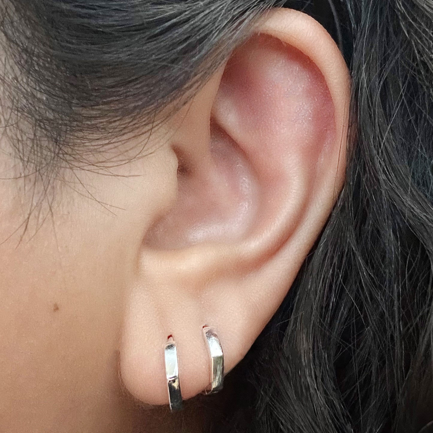 Model wearing two sterling silver hexagonal-shaped huggie hoop earrings. A larger earring is in her first lobe piercing.  The second earring is sitting snug against her lobe in her second piercing.
