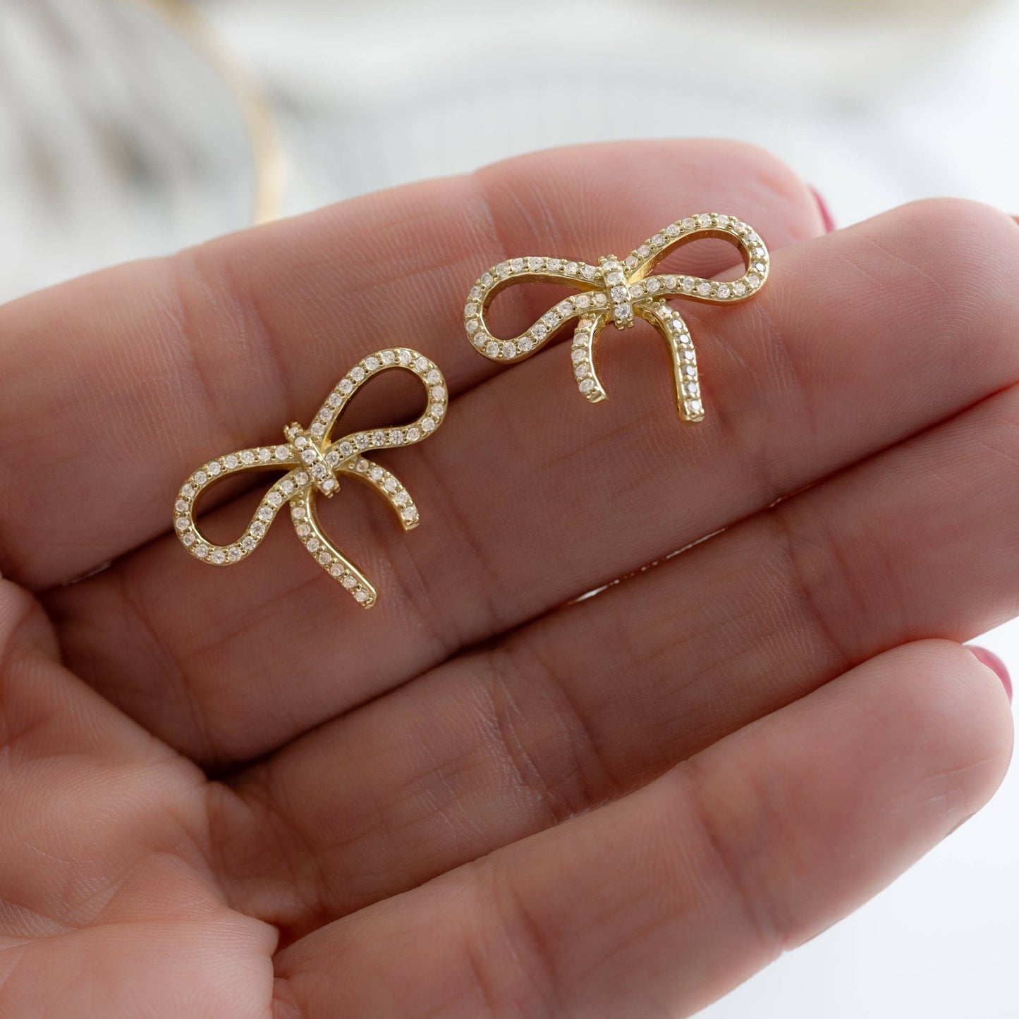 Gold-plated sterling silver stud earrings in the shape of a bow held between two fingers on a persons open hand. Each bow is lined with pave-set white cubic zirconia.