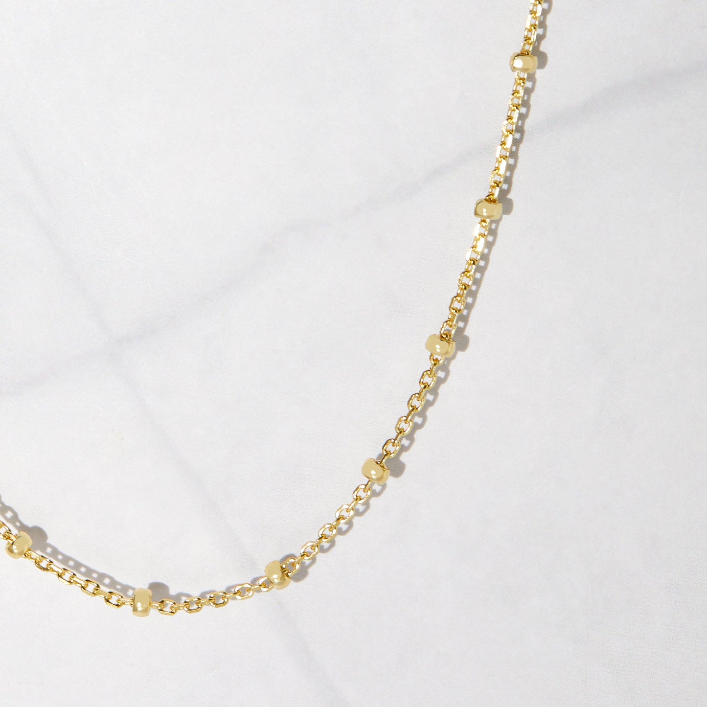 Close up of a dainty gold-plated sterling silver chain with gold beads hanging against a white stone background. The beads are separated by one inch of chain.