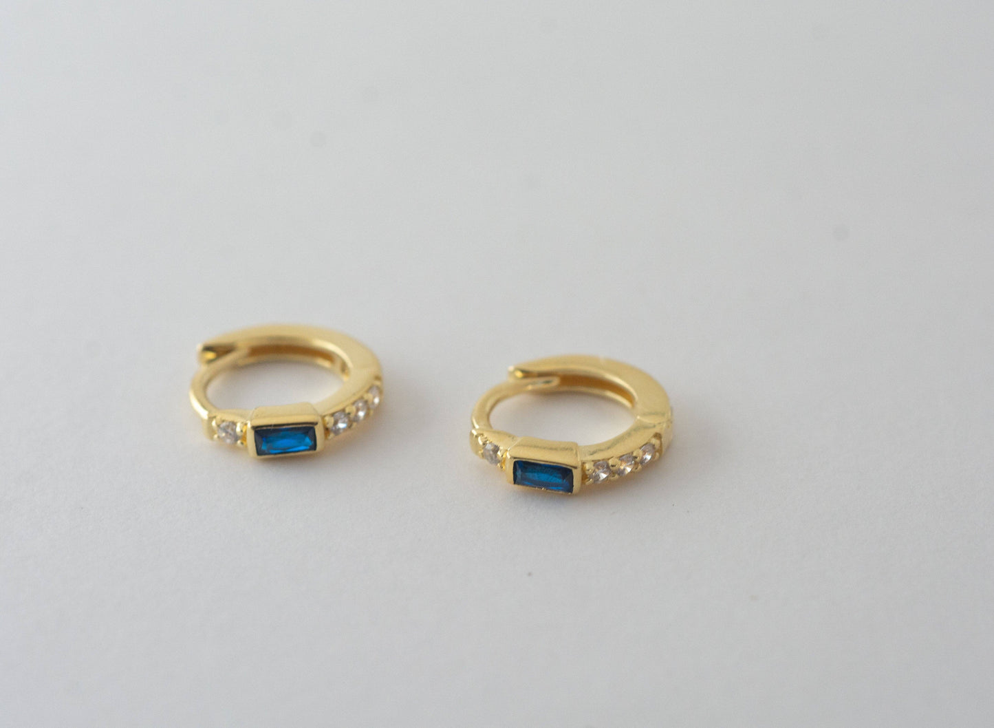 Top-down profile of a pair of small gold-plated huggie hoop earrings with a blue baguette-shaped cubic circonia. There is one white cubic zirconia above the blue stone and a line of 3 white cubic zirconias below. 