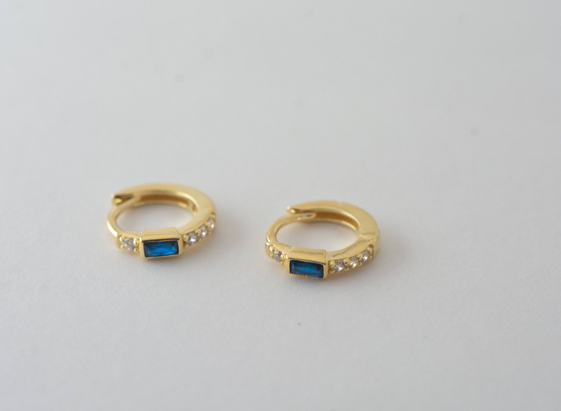 Top-down profile of a pair of small gold-plated huggie hoop earrings with a blue baguette-shaped cubic circonia. There is one white cubic zirconia above the blue stone and a line of 3 white cubic zirconias below. 