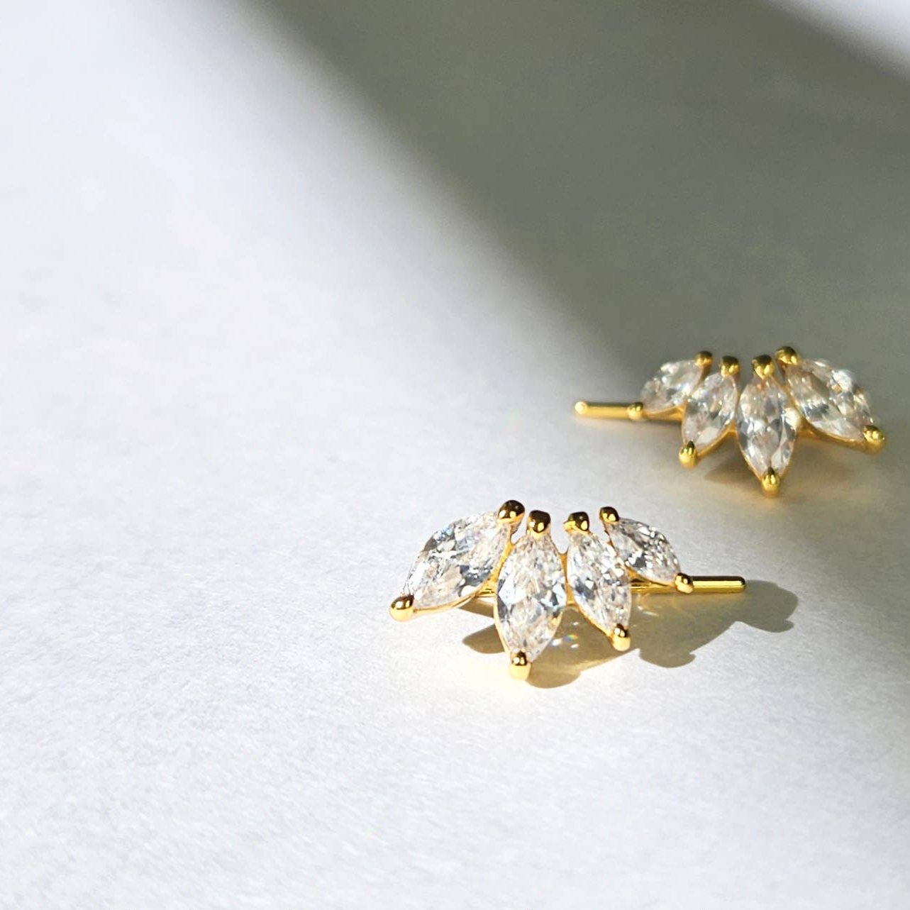 A pair of gold-plated sterling silver ear climbers sitting on a white stone surface. There are 4 marquis-cut white cubic zirconia set in a slight angle to create a shallow curve. 