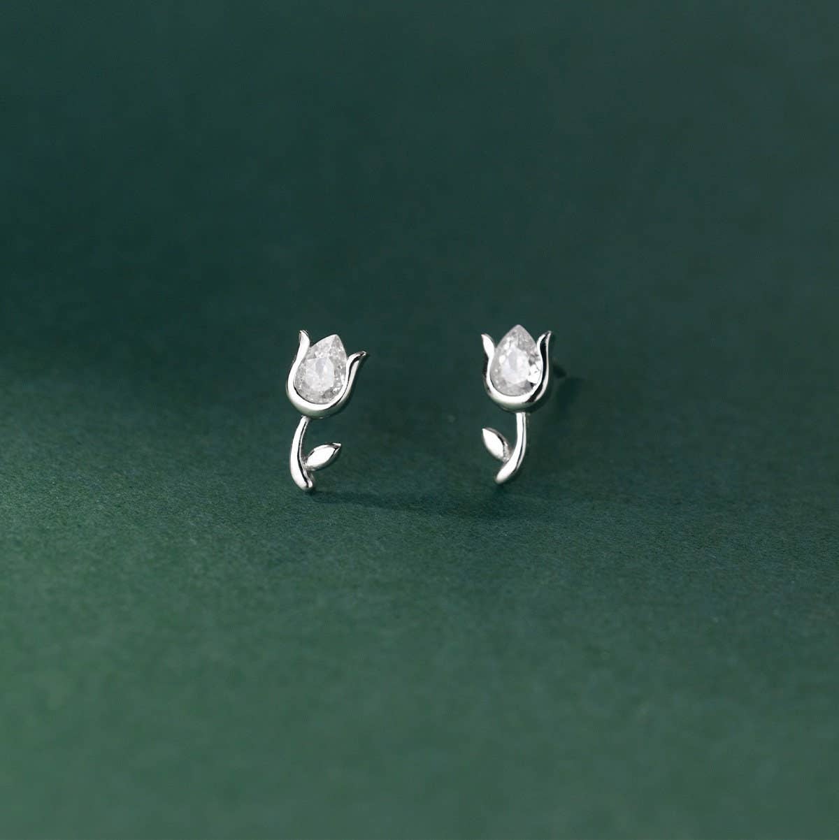 A pair of sterling silver tulip-shaped stud earrings against a green background. The earrings have a tear drop-shaped white cubic zirconia surrounded by sterling silver petals to form the flower. Each flower sits upon a sliver stem with a single silver leaf.  