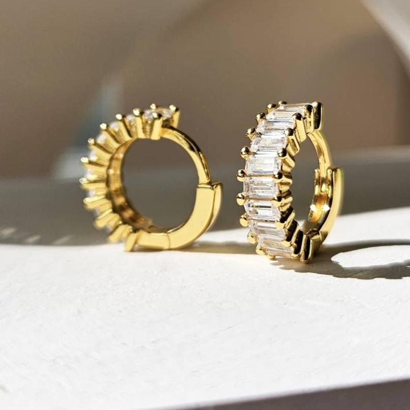 A pair of gold-plated sterling silver huggie hoop earrings. The front of the huggies are encrusted with a column of horizontal baguette shaped white cubic zirconia.  The earrings are sitting on a white stone counter. One earring is facing forward and the other is facing to the side showing the inside of the hoop.