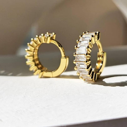 A pair of gold-plated sterling silver huggie hoop earrings. The front of the huggies are encrusted with a column of horizontal baguette shaped white cubic zirconia.  The earrings are sitting on a white stone counter. One earring is facing forward and the other is facing to the side showing the inside of the hoop.
