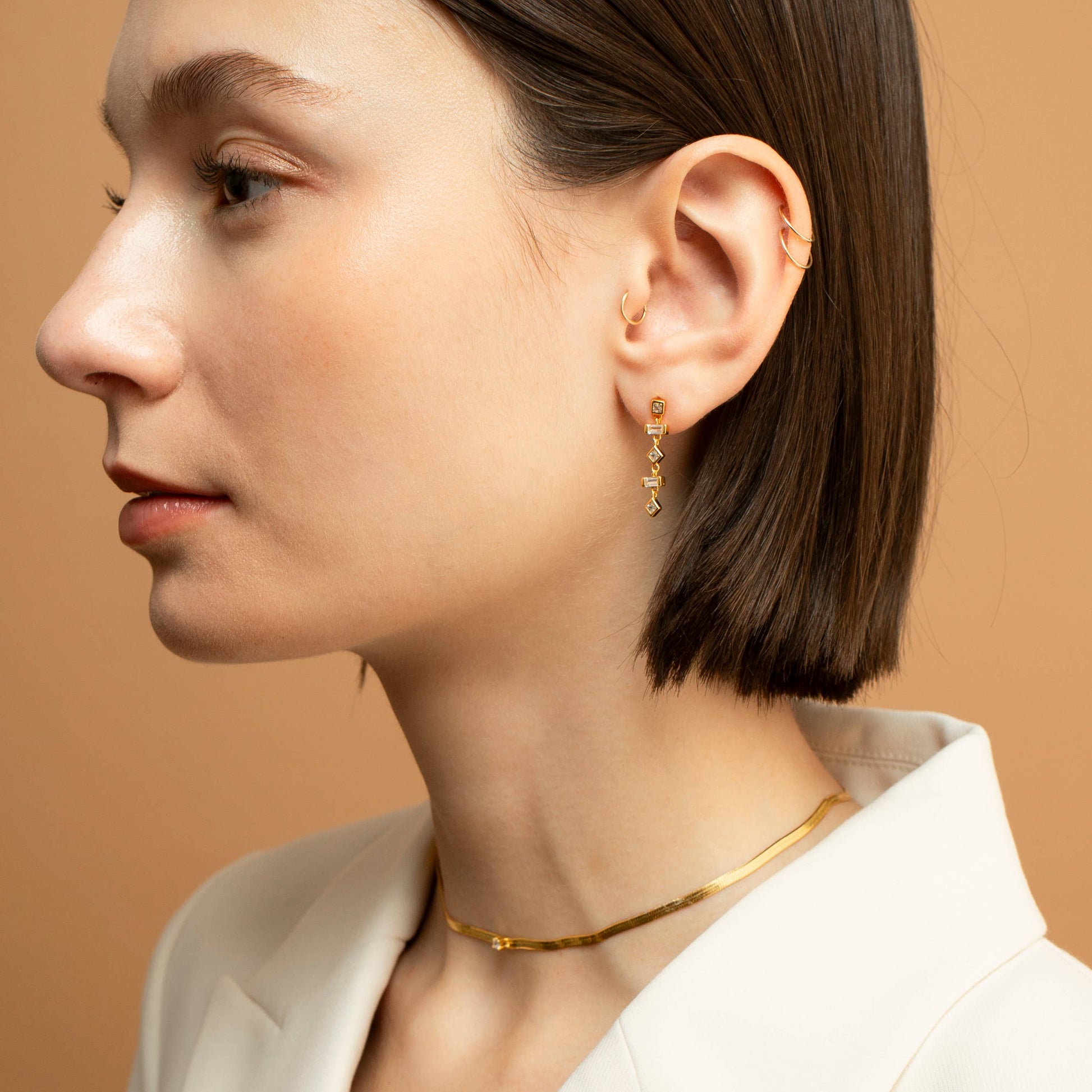 Model wearing a gold-plated sterling silver dangle earring with white cubic zirconia. There are 5 bead like elements that make up each earring. The top element is a square bezel set cubic zirconia. The second and fourth elements are baguette-shaped cubic zirconia. Elements 3 and 5 are diamond shaped cubic zirconia. 