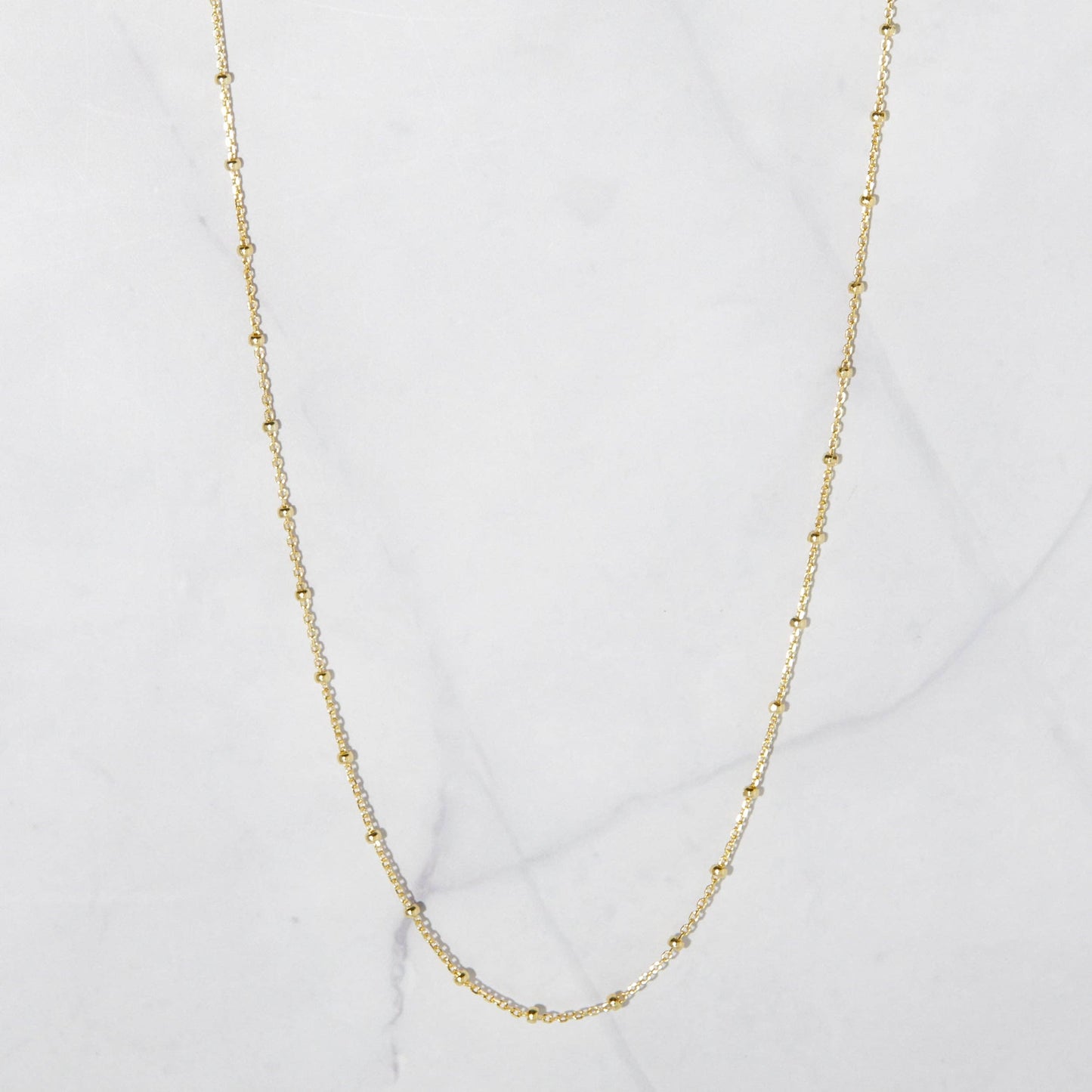 Dainty gold-plated sterling silver chain with gold beads hanging against a whote stone background. The beads are separated by one inch of chain. 