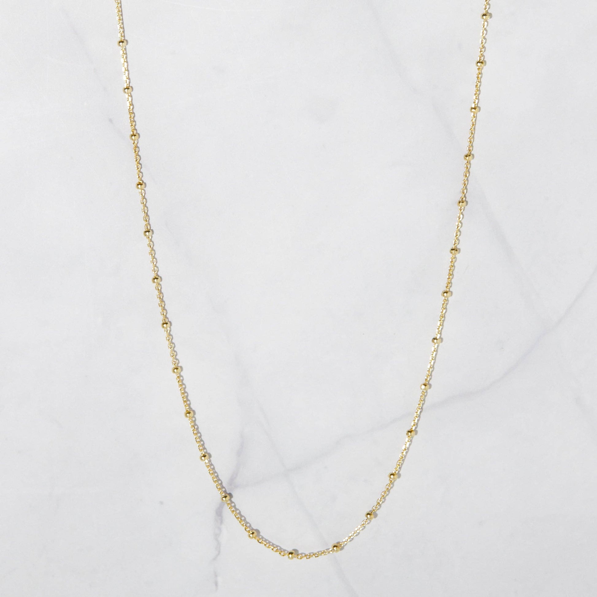 Dainty gold-plated sterling silver chain with gold beads hanging against a whote stone background. The beads are separated by one inch of chain. 