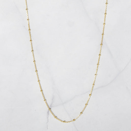Dainty gold-plated sterling silver chain with gold beads hanging against a whote stone background. The beads are separated by one inch of chain. 