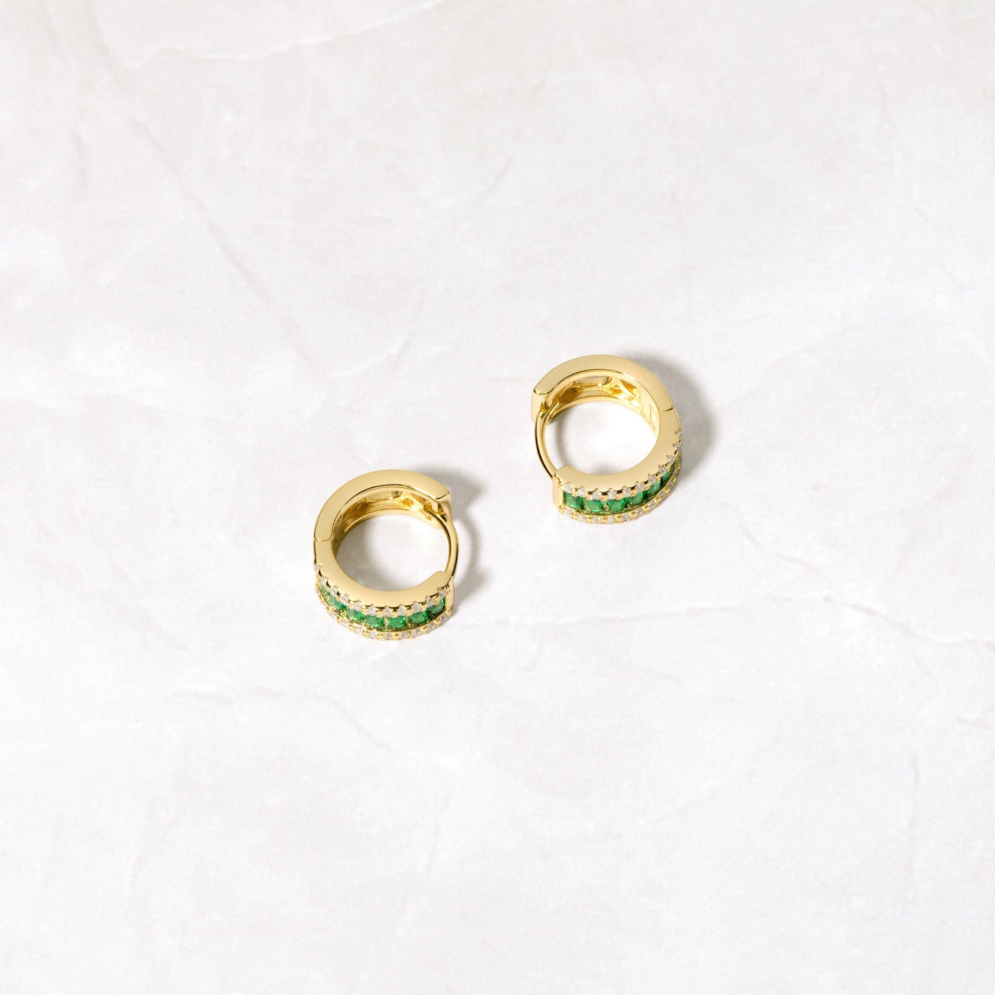 Side view of a pair of small gold-plated sterling silver huggie hoopssittign on a white stone surface. Each hugggie is set with a column of horizontal baguette cut green cubic zirconia. On either side of this column are small white cubic zirconia. 