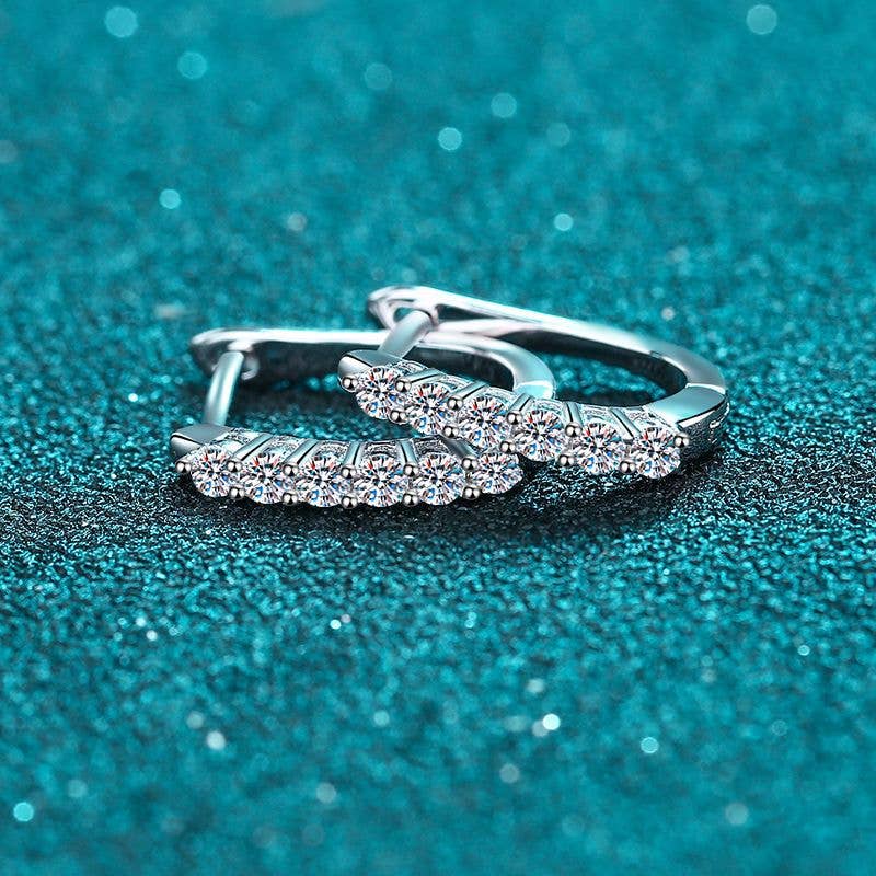 A pair of platinum-plated sterling silver oval hinged hoops laying on a glittery turquoise background. The front of the hoops are lined with 6 round-cut moissanite stones. The earrings are laying on top of each other.