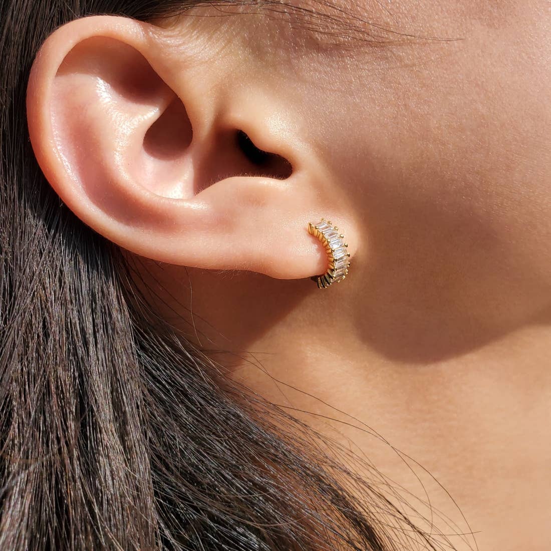 Model wearing a gold-plated sterling silver huggie hoop earring. The front of the huggies are encrusted with a column of horizontal baguette shaped white cubic zirconia. 