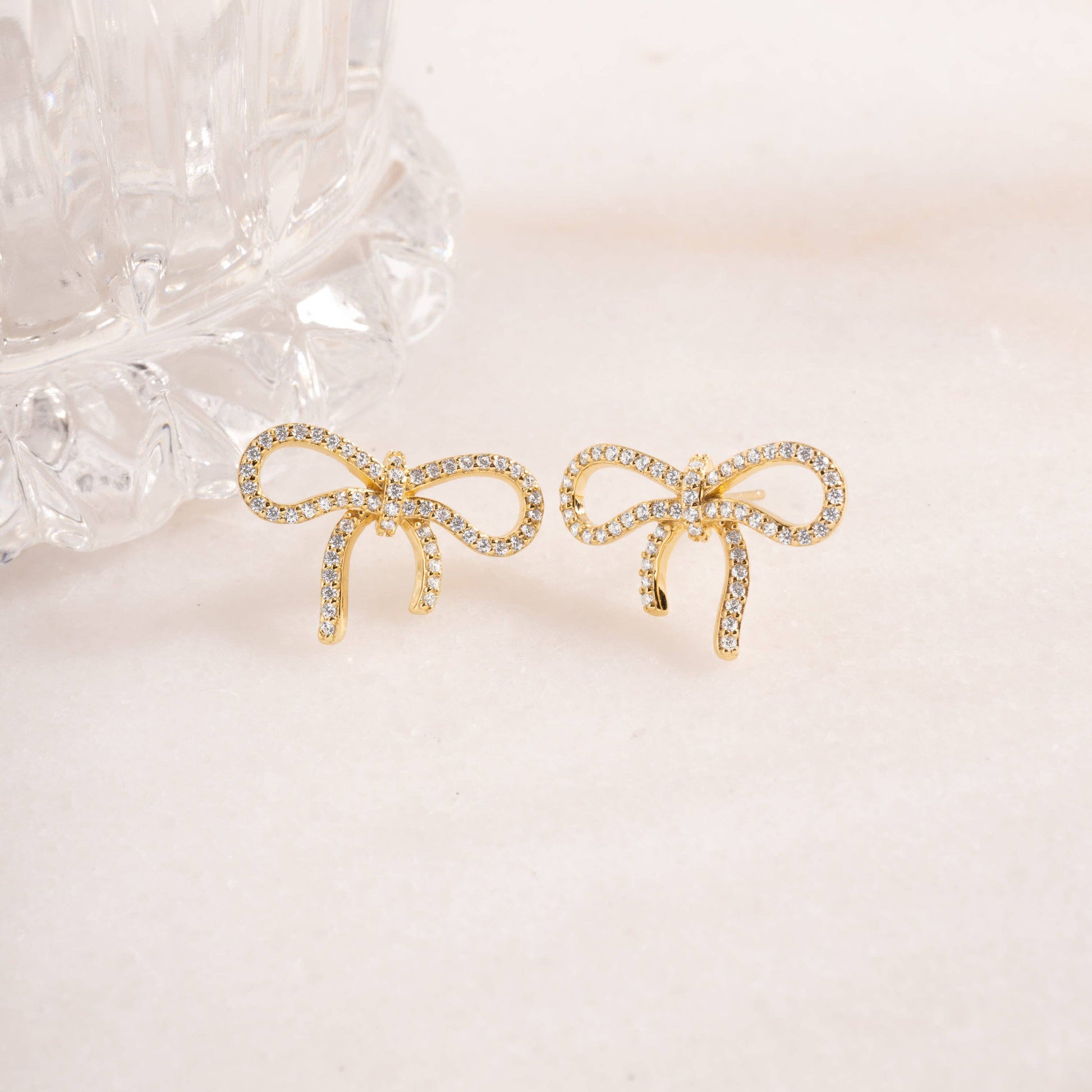 Gold-plated sterling silver stud earrings in the shape of a bow against a light pink background. Each bow is lined with pave-set white cubic zirconia.