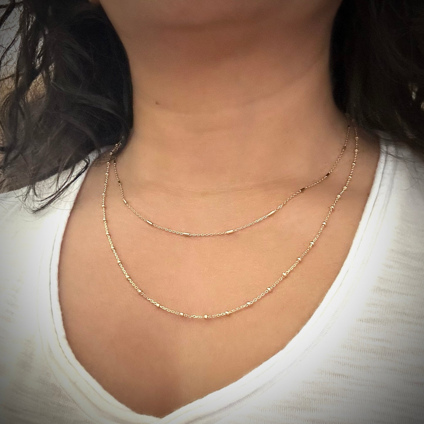 Model wearing a gold-plated sterling silver delicate chain necklace with elongated gold bead-like elements approximately every two inches. The model is also wearing a longer gold chain with small round beads placed an inch apart.
