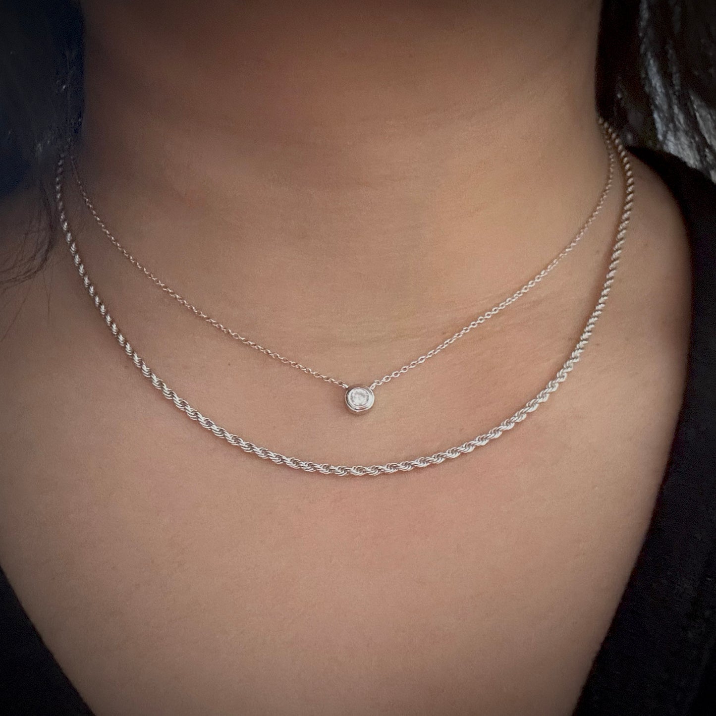 Model wearing a sterling silver delicate chain necklace with bezel set white cubic zirconia pendant connected to the chain. The model has layered this necklace with a sterling silver rope chain necklace.
