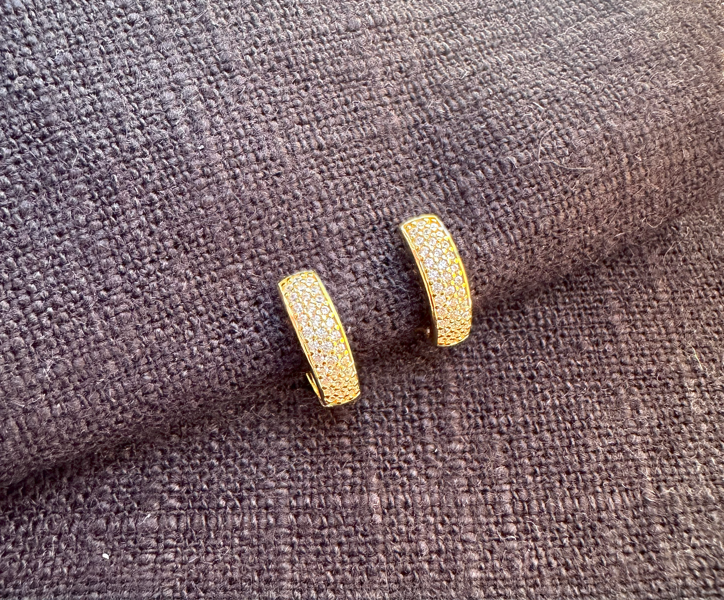 A pair of gold-plated sterling silver huggie hoop earrings set against black linen. The earrings have 3 columns of white cubic zirconia set in a pave style.