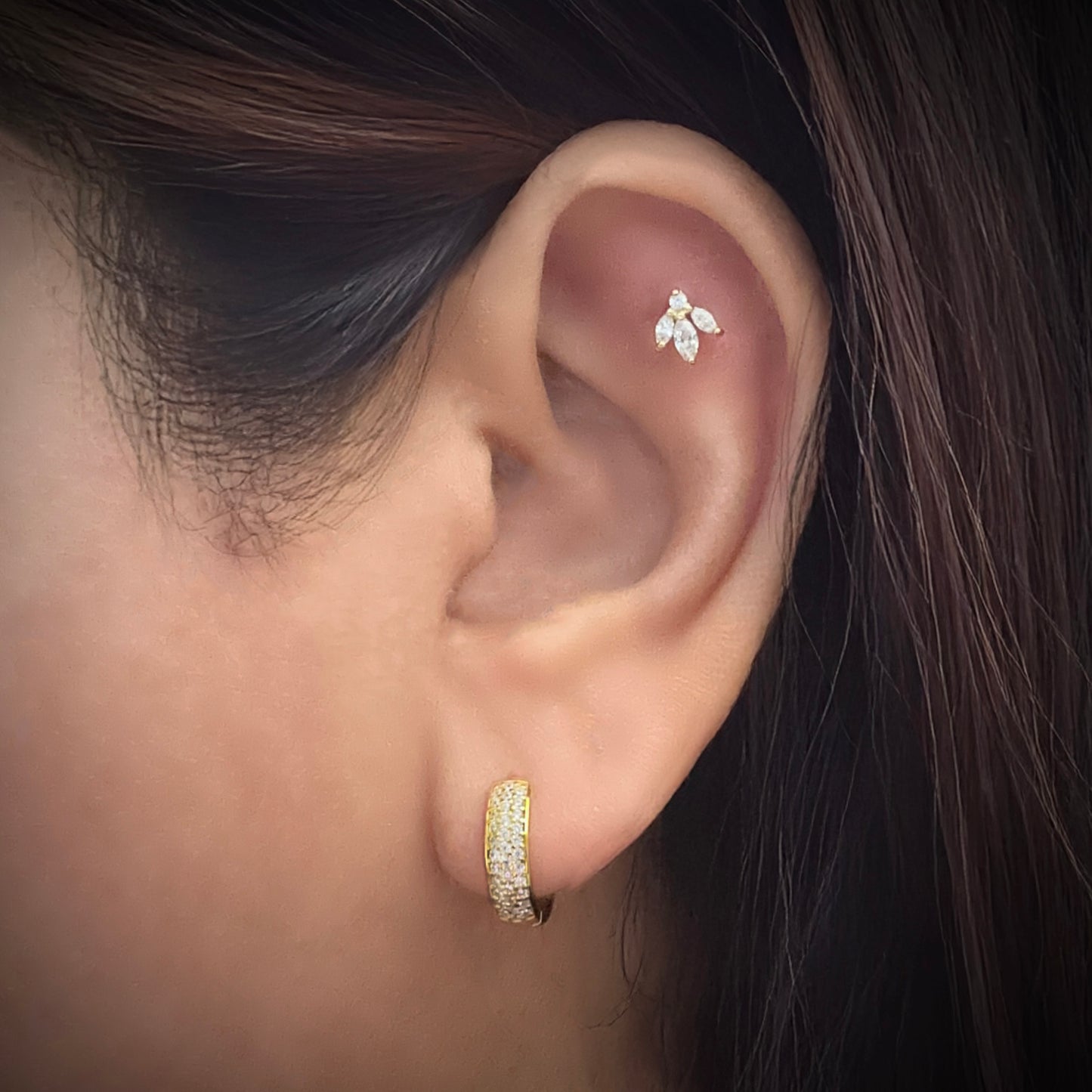 Model wearing a gold-plated sterling silver huggie hoop earrings. The earring has 3 columns of white cubic zirconia set in a pave style. The model is also wearing a flower-shaped white cubic zirconia stud with 3 petals set below a round white zirconia stone in the flat part of her ear just below the upper helix. 