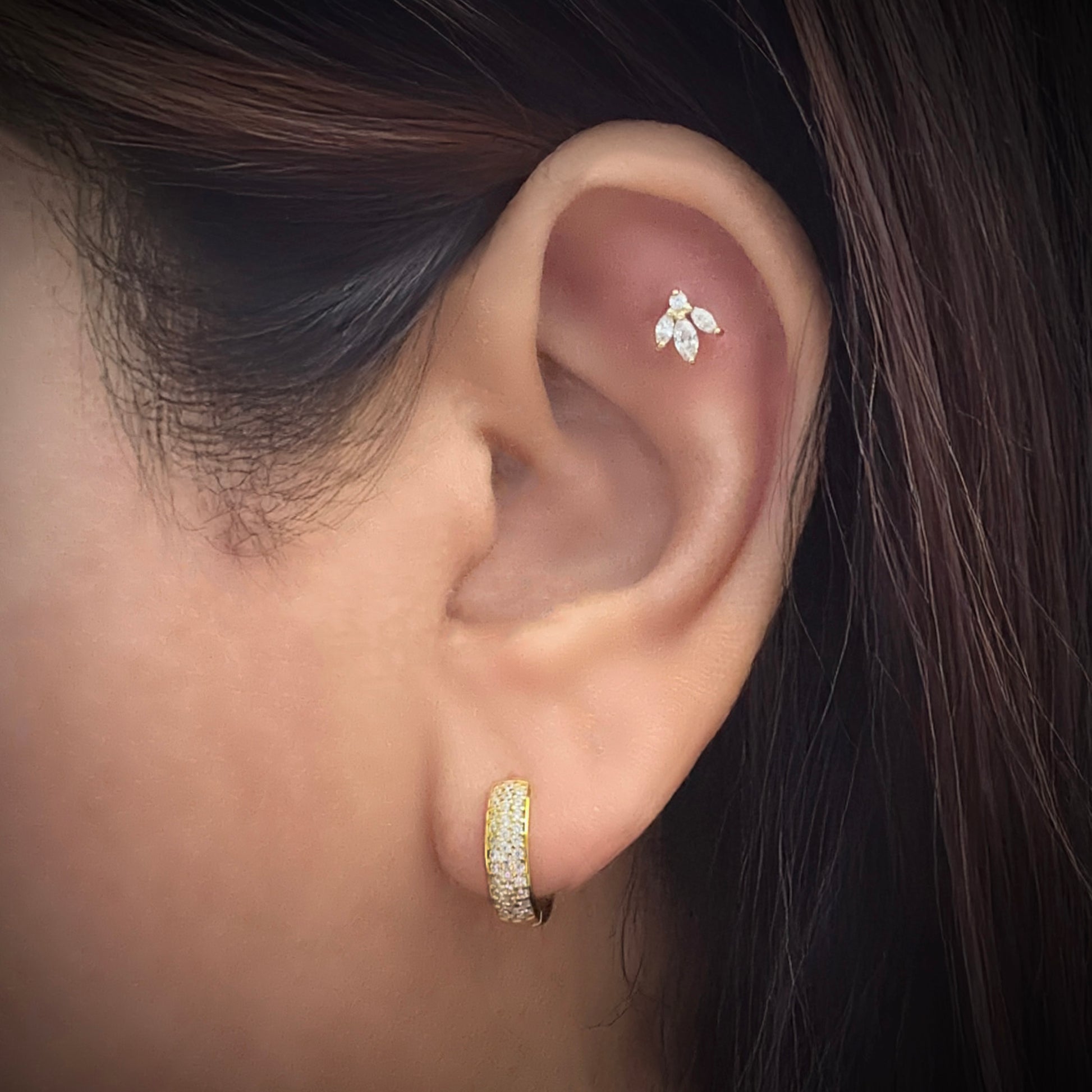 Model wearing a gold-plated sterling silver huggie hoop earrings. The earring has 3 columns of white cubic zirconia set in a pave style. The model is also wearing a flower-shaped white cubic zirconia stud with 3 petals set below a round white zirconia stone in the flat part of her ear just below the upper helix. 