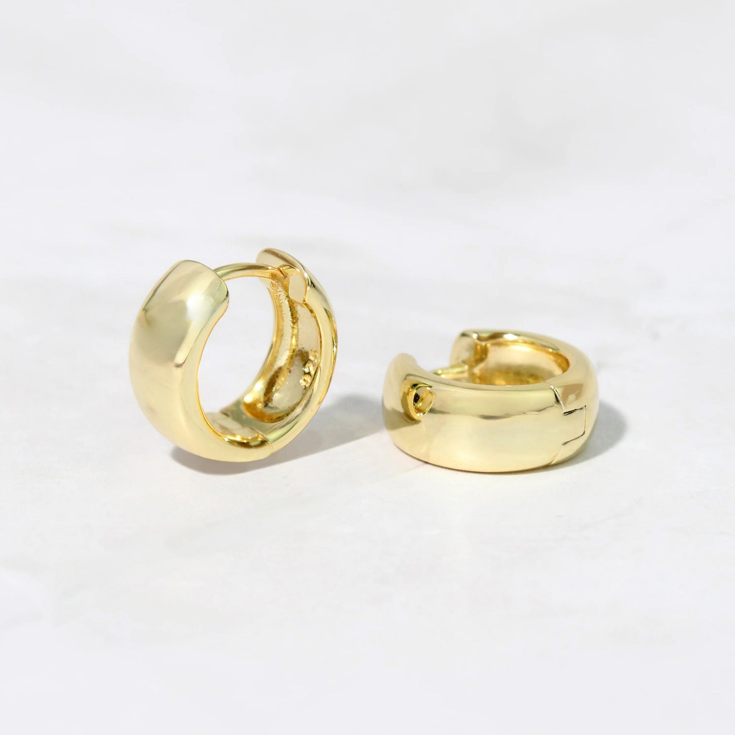 A pair of gold-plated sterling silver chunky huggie hoop earrings. One earring is standing up showing the inside of the hoop. 925 is stamped on the inside. The other earring is laying on its side. The earrings are on a gray background.