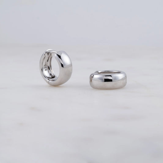A pair of sterling silver chunky huggie hoop earrings. One earring is standing up showing the inside of the hoop. 925 is stamped on the inside. The other earring is laying on its side. The earrings are on a gray background.