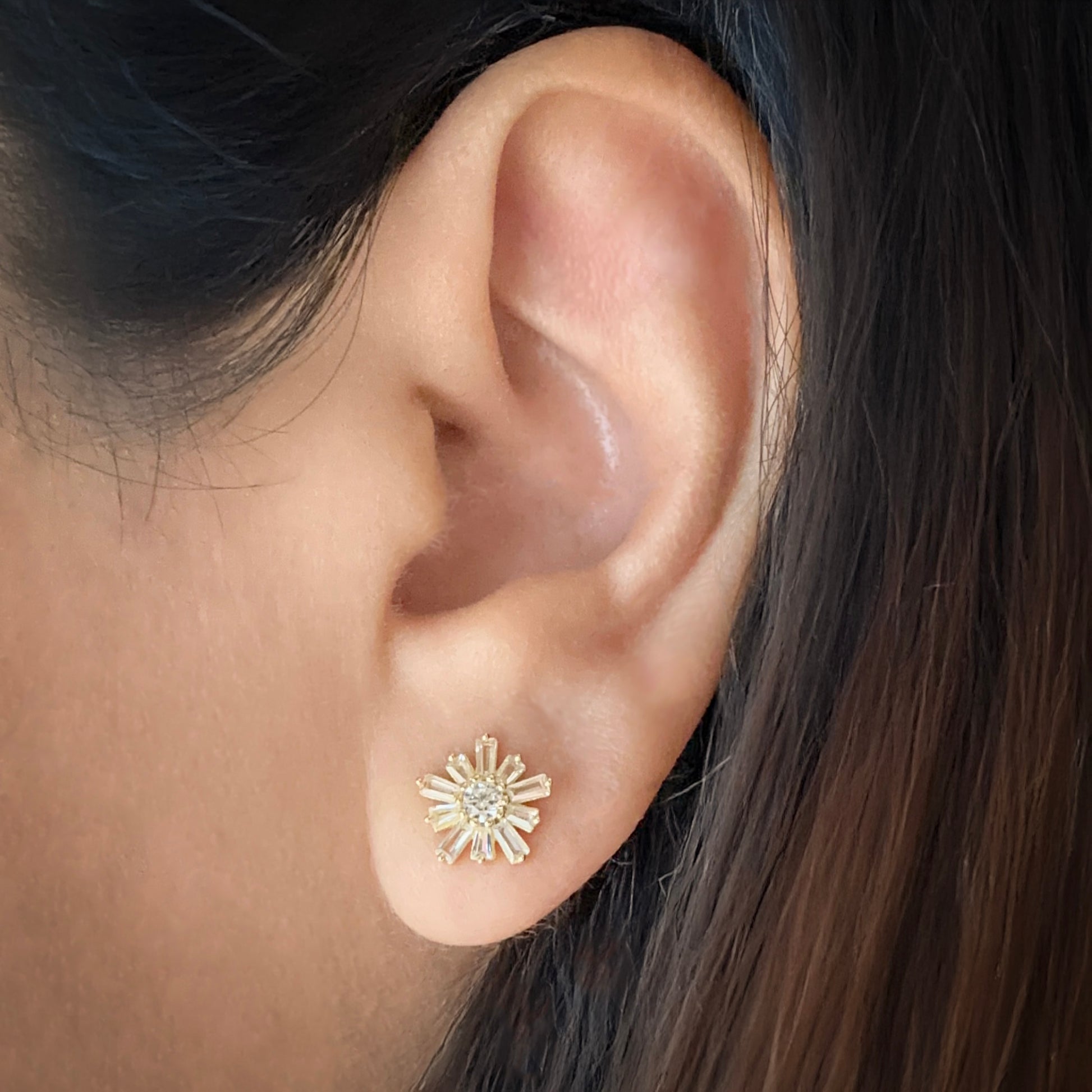 Model wearing Sterling silver flower-shaped studs earrings with 10 petals and a circular stone in the center. The image focuses on one ear with the earring in a traditional lobe piercing. Each petal is made of white cubic zirconia in a baguette shape. The center stone is white cubic zirconia. 