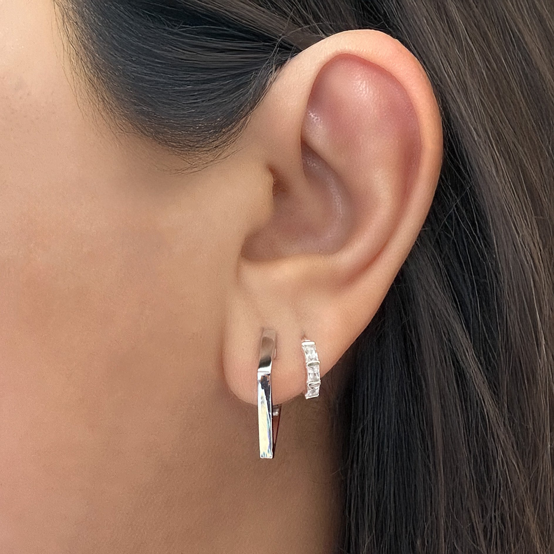 Model wearing a sterling silver kite-shaped hoop earring. The model is also wearing a small silver huggie hoop with baguette-cut white cubic zirconia in her second lobe piercing.