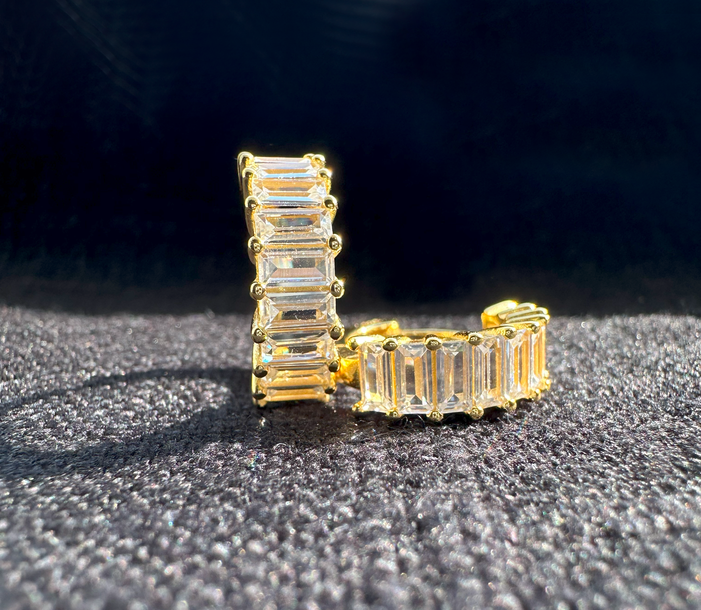 A pair of gold-plated sterling silver huggie hoop earrings. The front of the huggies are encrusted with a column of horizontal baguette shaped white cubic zirconia.  The earrings are sitting on black linen. One earring is facing forward and the other is laying on it's side.
