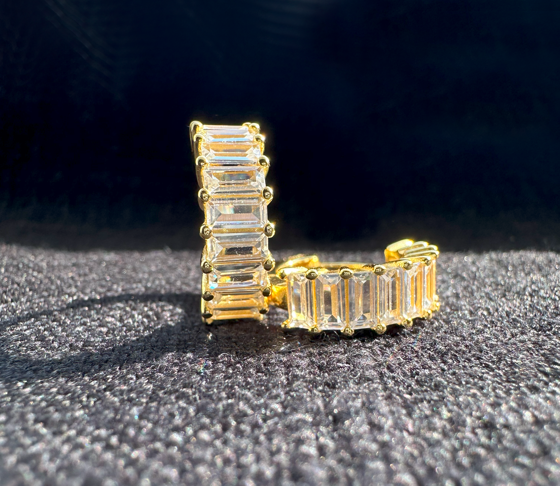 A pair of gold-plated sterling silver huggie hoop earrings. The front of the huggies are encrusted with a column of horizontal baguette shaped white cubic zirconia.  The earrings are sitting on black linen. One earring is facing forward and the other is laying on it's side.