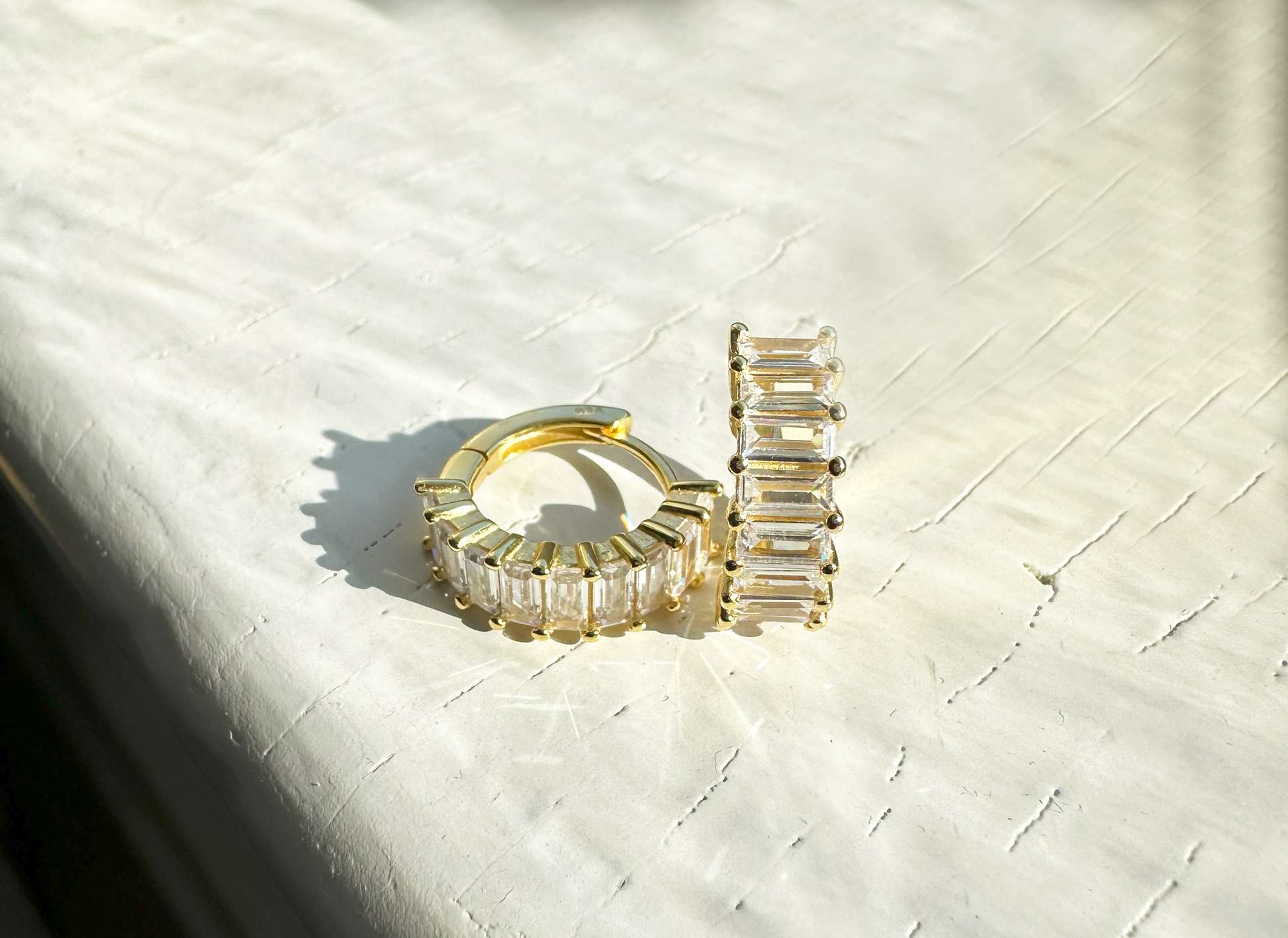 A pair of gold-plated sterling silver huggie hoop earrings. The front of the huggies are encrusted with a column of horizontal baguette shaped white cubic zirconia.  The earrings are sitting on a white, wooden counter. One earring is facing forward and the other is facing to the side showing the inside of the hoop.