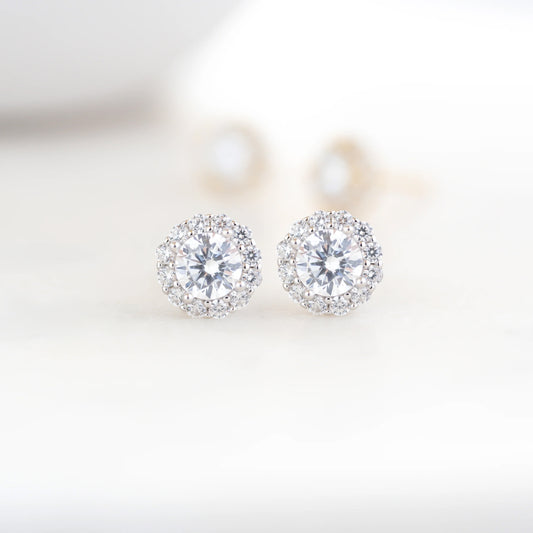 Sterling silver white cubic zirconia stud earrings sitting on a white counter. The earrings have 12 small stones surround a larger round stone in the center. Faded in the background are a pair of the same studs in gold-plated sterling silver. 