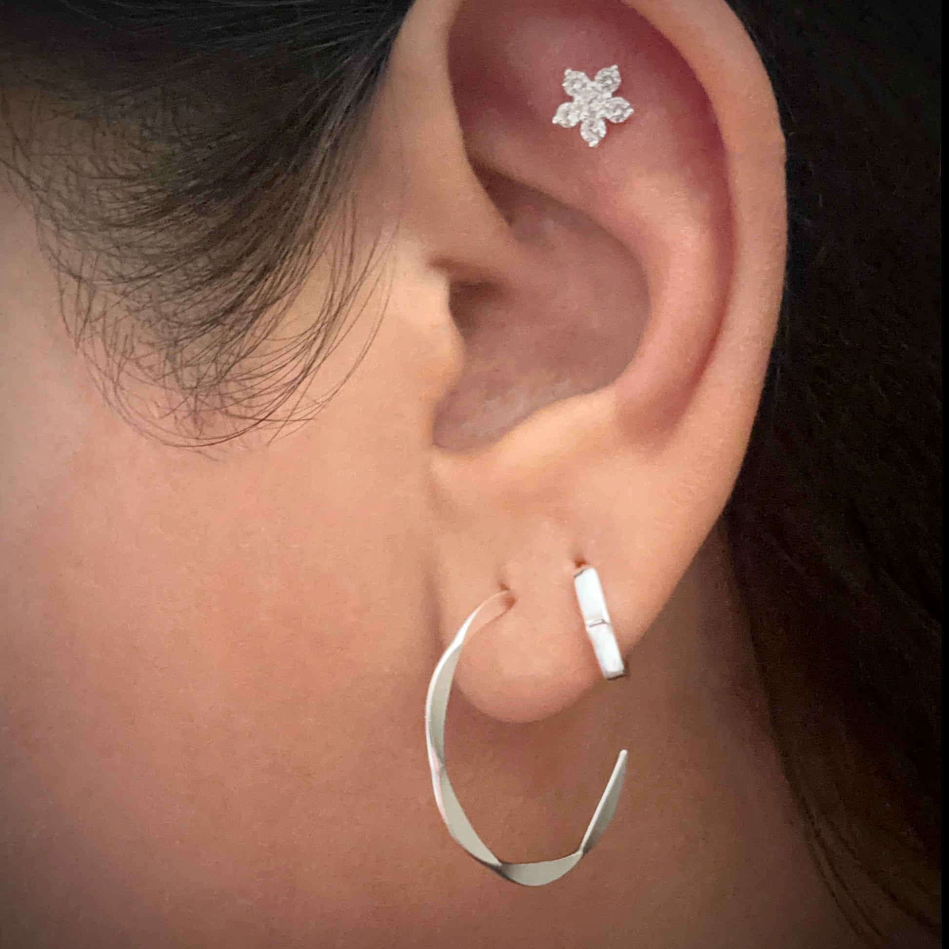 Model wearing a sterling silver hexagonal-shaped huggie hoop earring in her second lobe piercing. She is also wearing a silver demi hoop in her first lobe piercing and a flower-shaped white cubic zirconia stud in the flat part of her ear just under the upper helix. 
