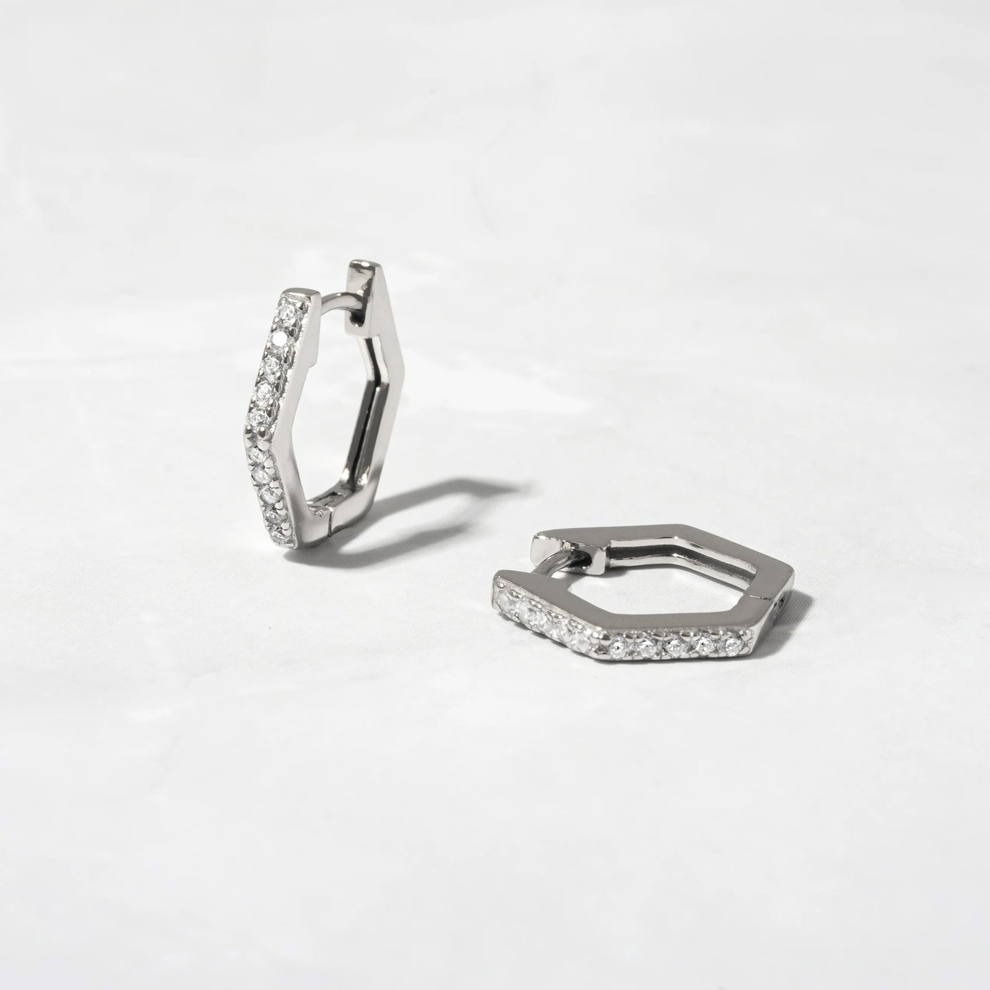 A pair of sterling silver hexagonal-shaped huggie earringsSitting on a stone background. The earrings are lined with round white cubic zirconias. One earring is standing up showing the front and side of the earring and the shape. The other earring is laying on the surface. 