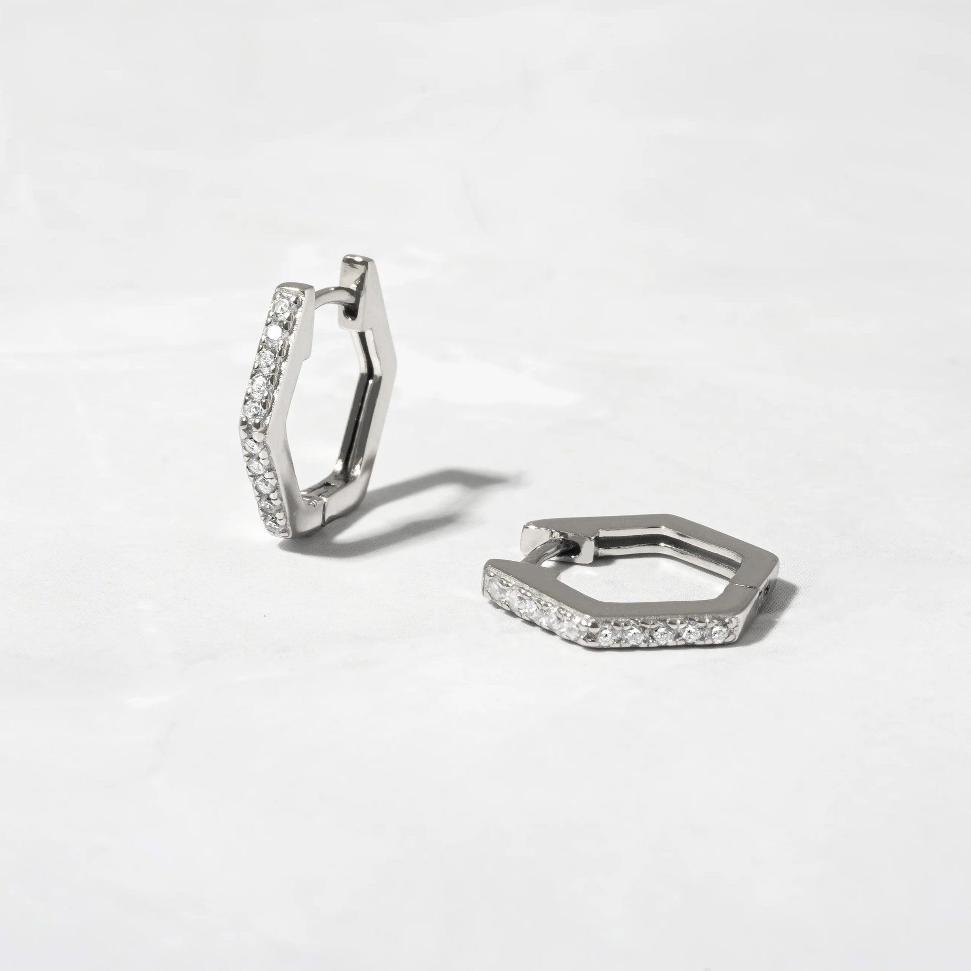A pair of sterling silver hexagonal-shaped huggie earringsSitting on a stone background. The earrings are lined with round white cubic zirconias. One earring is standing up showing the front and side of the earring and the shape. The other earring is laying on the surface. 