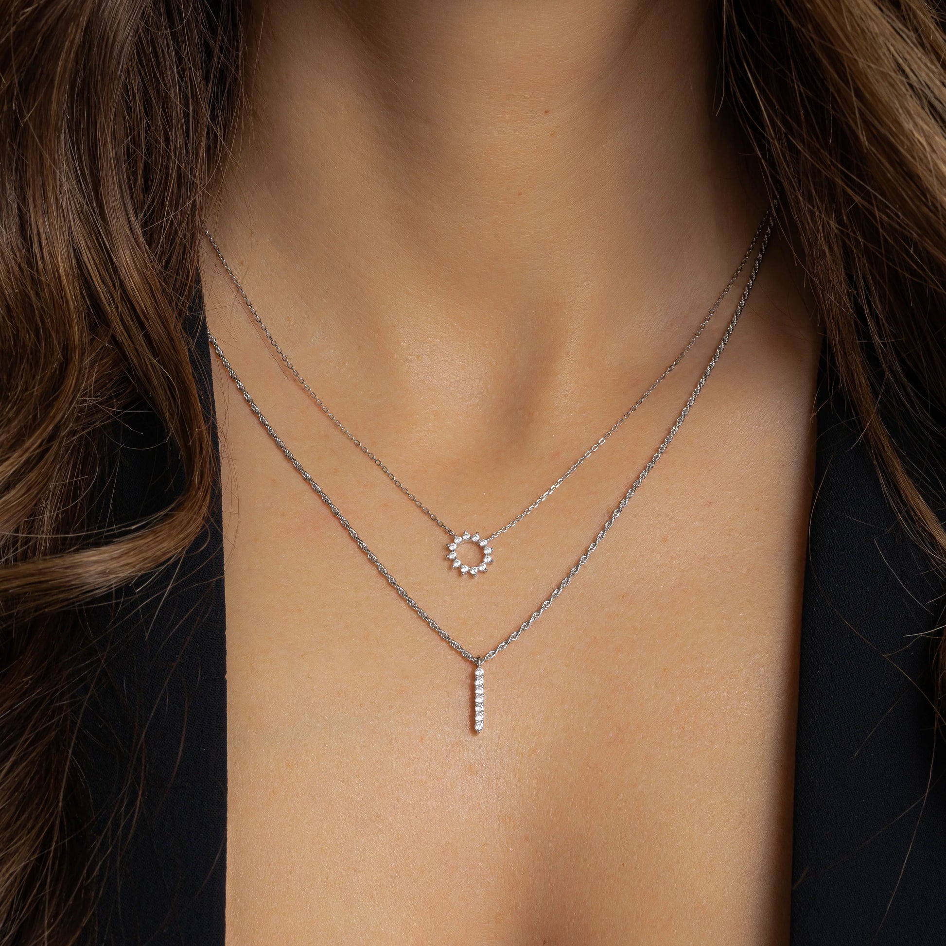 Model wearing a delicate sterling silver necklace with a circular pendant. The pendant is an open circle with 12 white cubic zirconia stones set around the circumference in the shape of the sun. The model is also wearing a rope chain necklace that sits below the sunshine necklace.