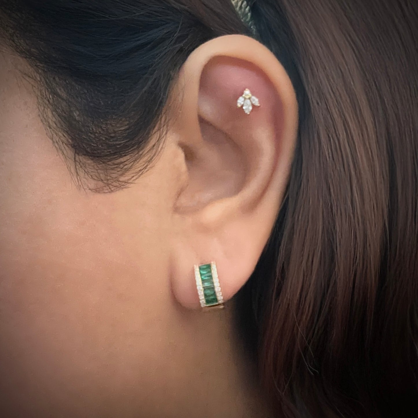 Model wearing a small gold-plated sterling silver huggie hoop. The hugggie is set with a column of horizontal baguette cut green cubic zirconia. On either side of this column are small white cubic zirconia. 