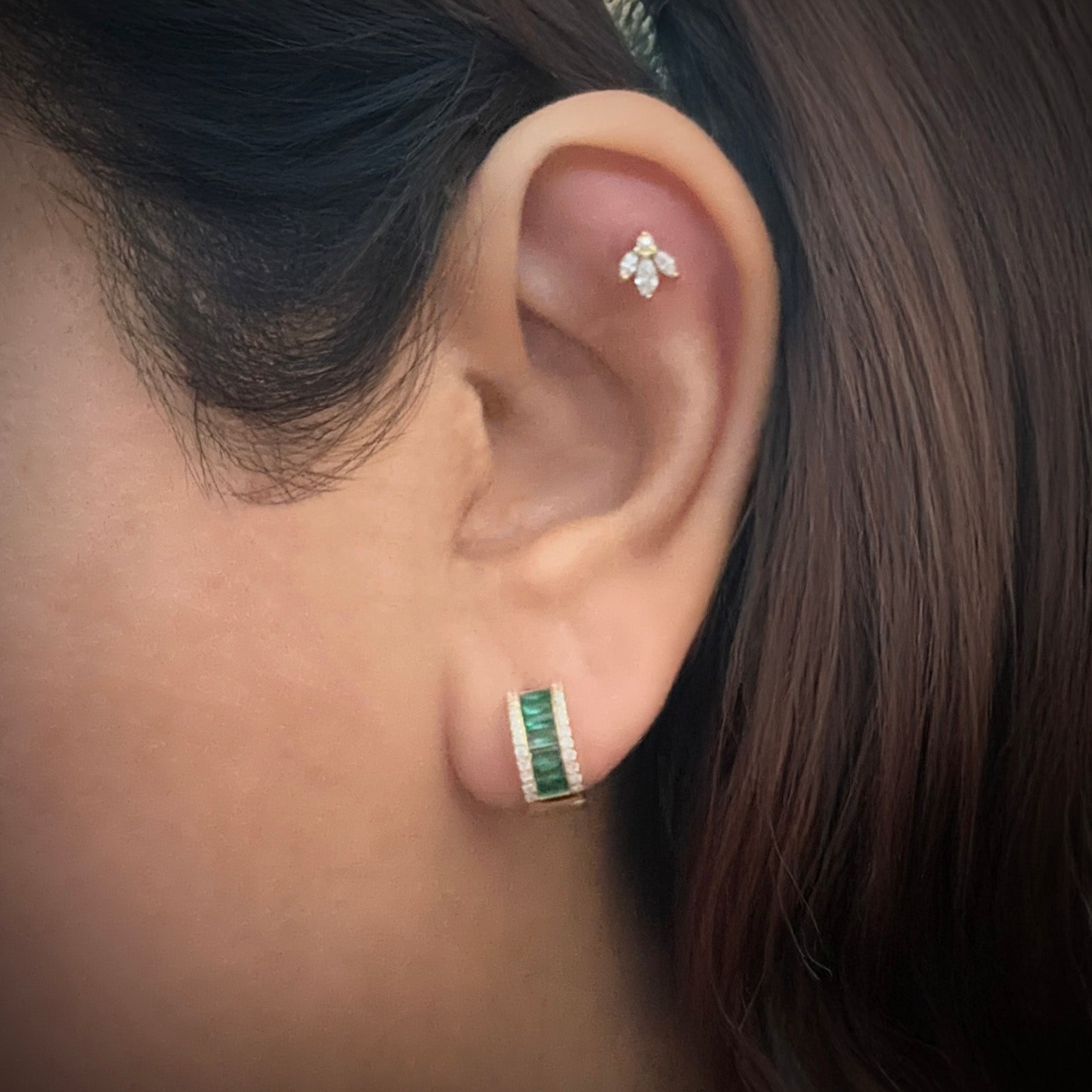 Model wearing a small gold-plated sterling silver huggie hoop. The hugggie is set with a column of horizontal baguette cut green cubic zirconia. On either side of this column are small white cubic zirconia. 