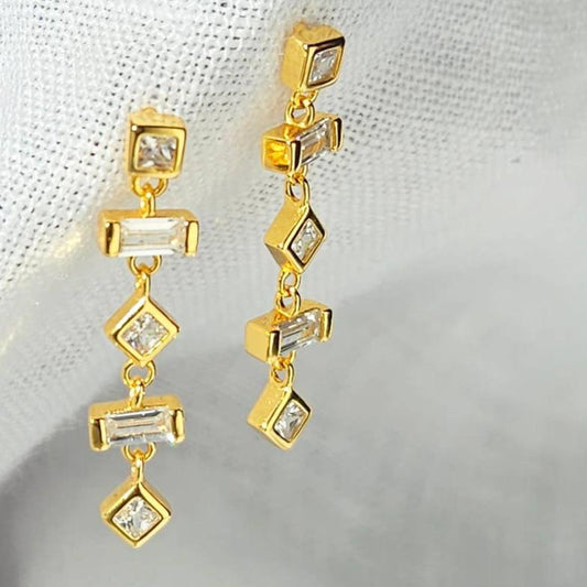 Pair of gold-plated sterling silver dangle earrings with white cubic zirconia. There are 5 bead like elements that make up each earring. The top element is a square bezel set cubic zirconia. The second and fourth elements are baguette-shaped cubic zirconia. Elements 3 and 5 are diamond shaped cubic zirconia. The earrings are dangling from white linen fabric.