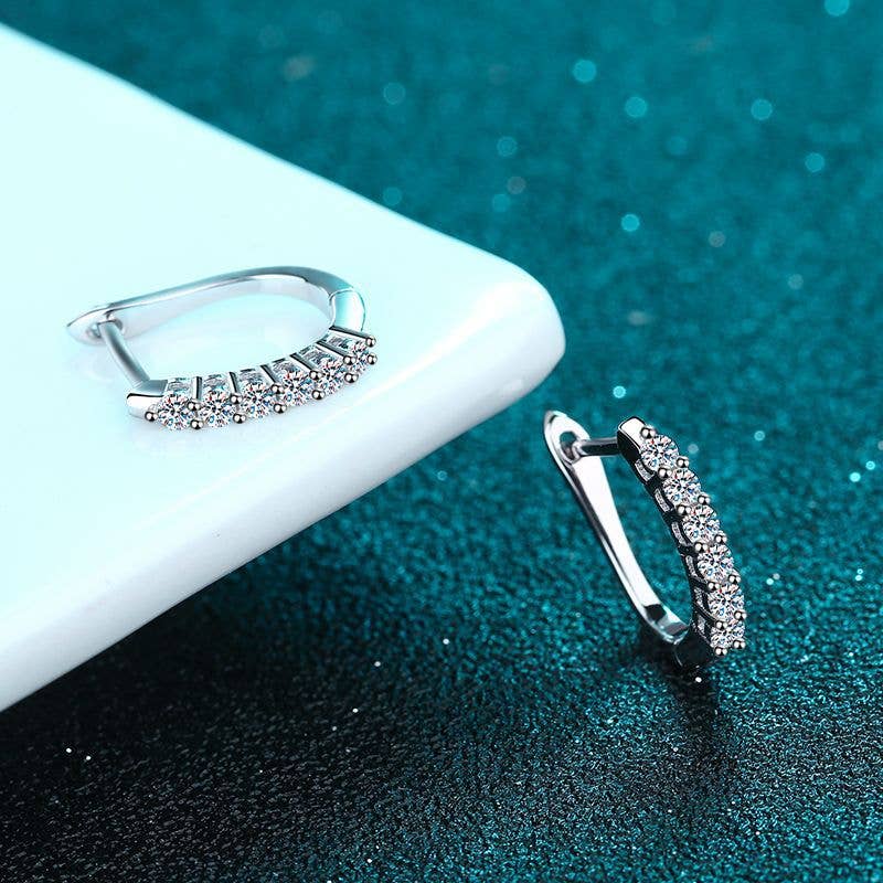 A pair of platinum-plated sterling silver oval hinged hoops sitting. The front of the hoops are lined with 6 round-cut moissanite stones. One earring is laying on a white shelf. The other earring is standing on a glittery turquoise background. 