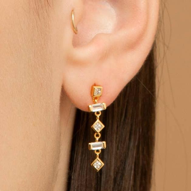 Close-up of a model wearing a gold-plated sterling silver dangle earring with white cubic zirconia. There are 5 bead like elements that make up each earring. The top element is a square bezel set cubic zirconia. The second and fourth elements are baguette-shaped cubic zirconia. Elements 3 and 5 are diamond shaped cubic zirconia. 