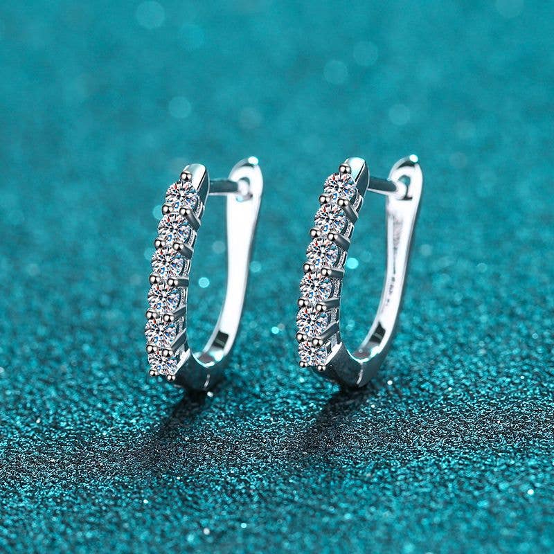 A pair of platinum-plated sterling silver oval hinged hoops sitting on a glittery turquoise background. The front of the hoops are lined with 6 round-cut moissanite stones. The earrings are slightly facing to the left showing the hinge and clasp mechanism.