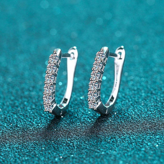 A pair of platinum-plated sterling silver oval hinged hoops sitting on a glittery turquoise background. The front of the hoops are lined with 6 round-cut moissanite stones. The earrings are slightly facing to the left showing the hinge and clasp mechanism.