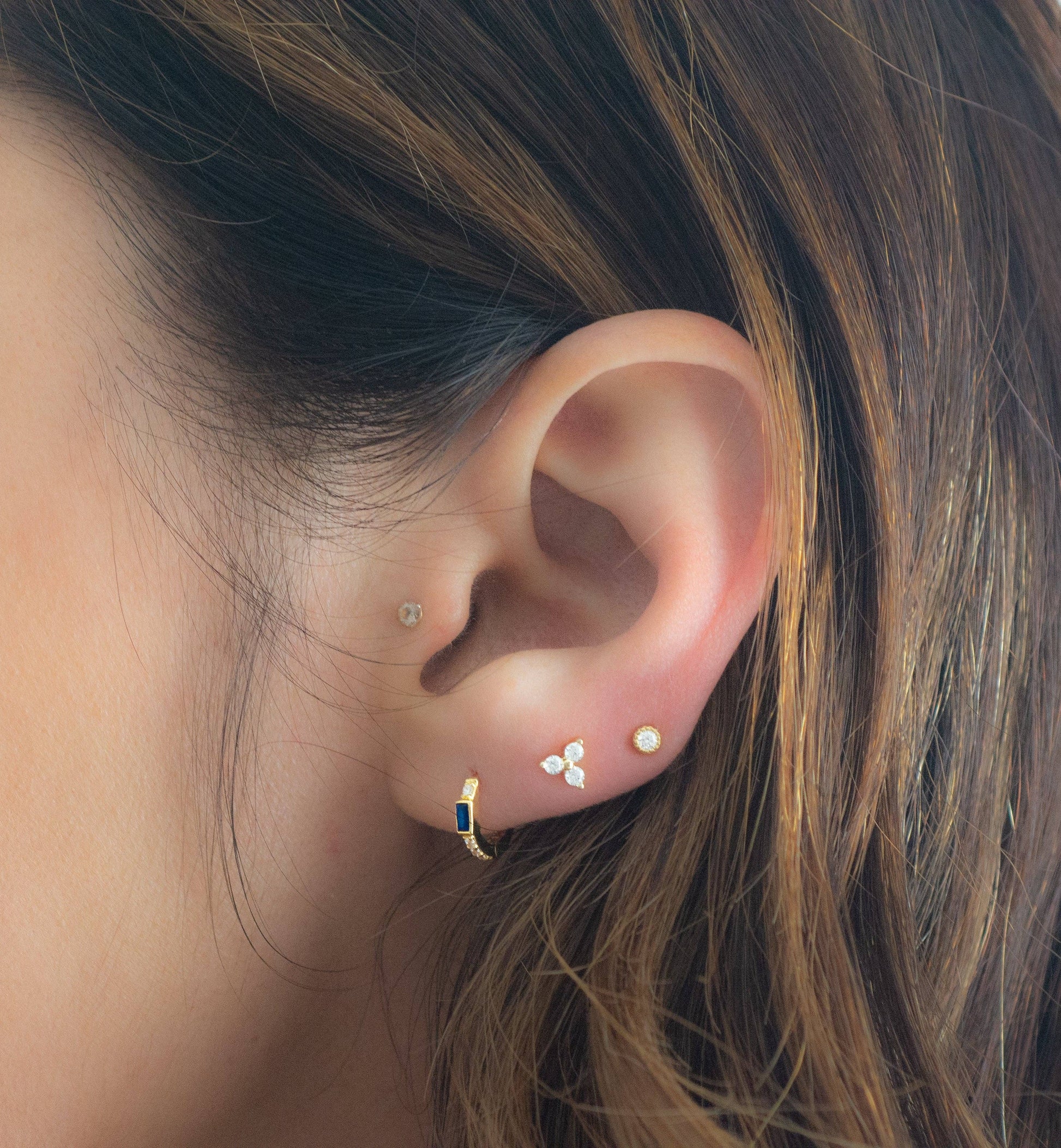 Model wearing a small gold-plated huggie hoop earring with a blue baguette-shaped cubic circonia. There is one white cubic zirconia above the blue stone and a line of 3 white cubic zirconias below. 